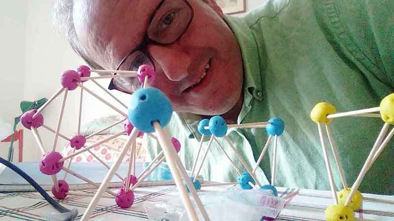 3-dimensional geometric structures built by joining toothpicks with balls of clay of different colors. The author of the piece is on the frame, behind the structures, looking at the camera.