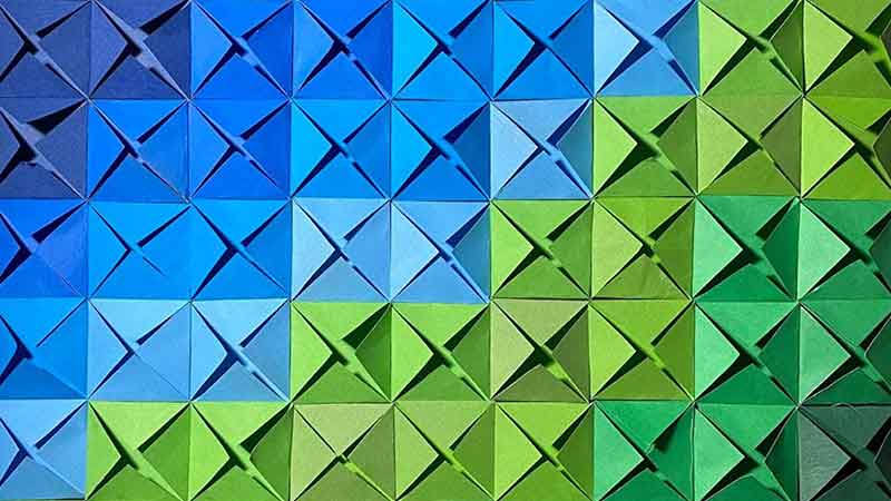 A rectangular grid composed of folded paper squares, arranged to form a color gradient. Each square forms a small 3D, pyramid-like shape, creating a textured, geometric pattern against a dark background.