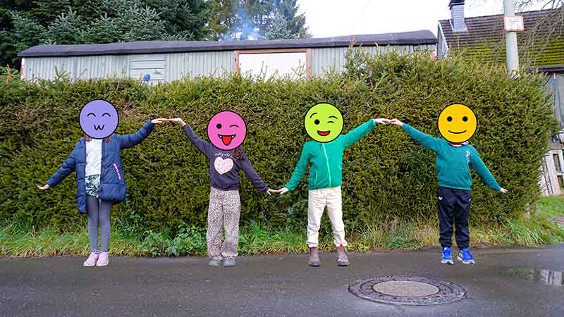 Four children stand hand in hand outside on a village street. Their hands are alternatively held up or down so their arms form a sine wave-like pattern.