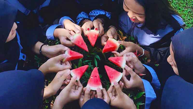 Eight people stand or sit in a circle, some wearing head coverings. Each person holds a small triangular slice of watermelon (one eight of a circle). The slices are arranged so that their tips point toward the center.