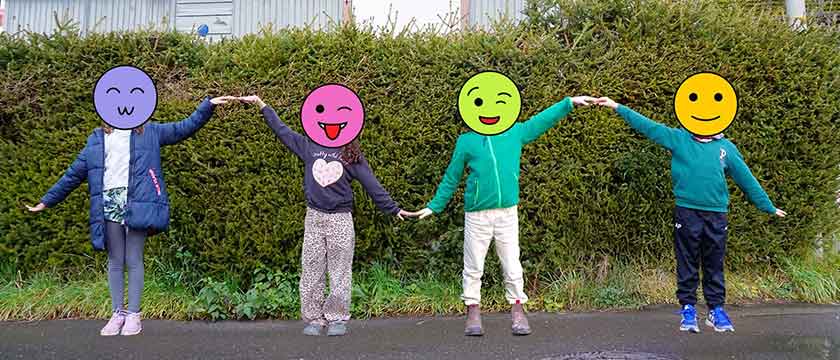 Four children stand hand in hand outside on a village street. Their hands are alternatively held up or down so their arms form a sine wave-like pattern.